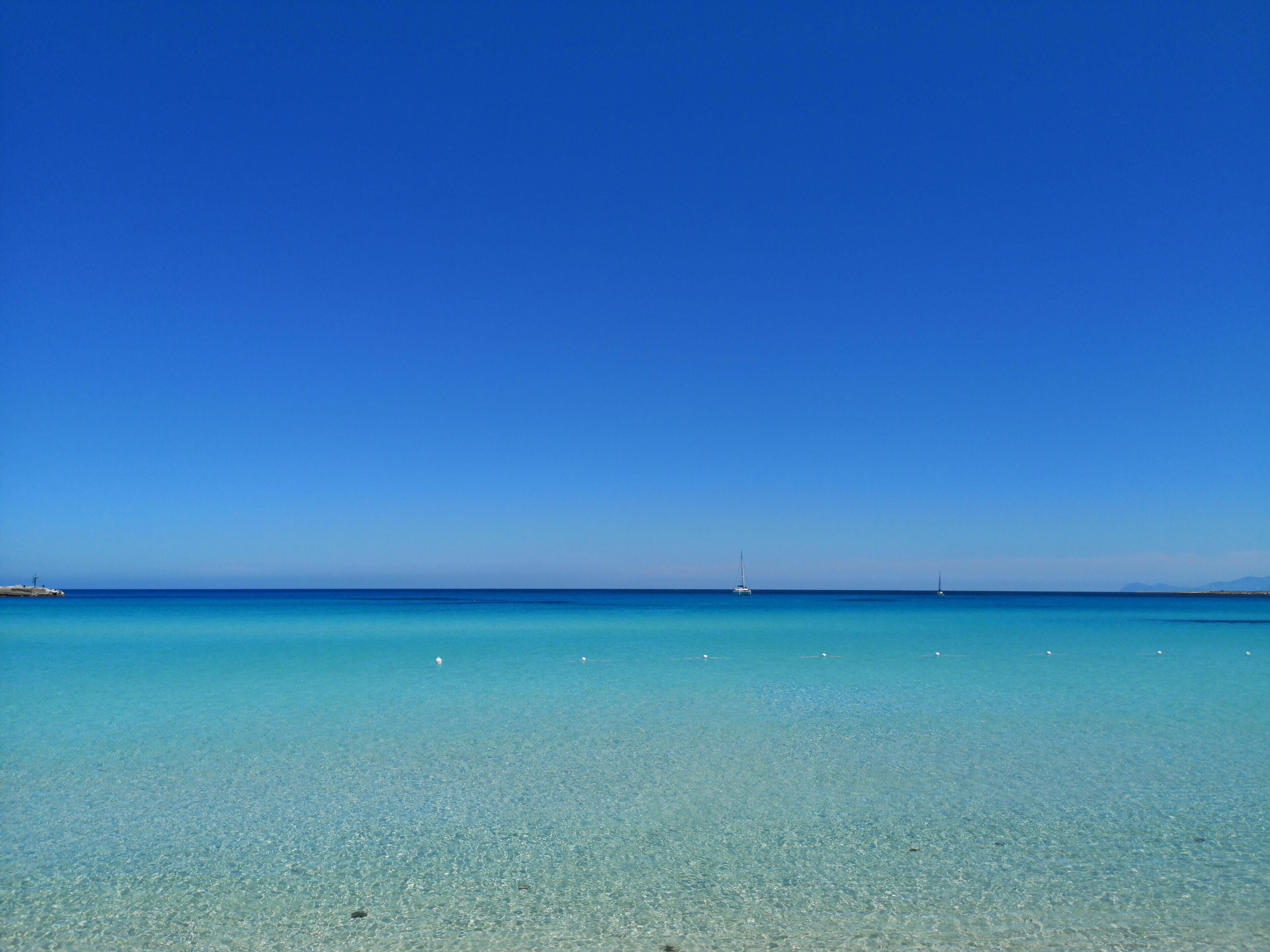 A clear blue sea with a distant sailboat and a rocky outcrop on the left.