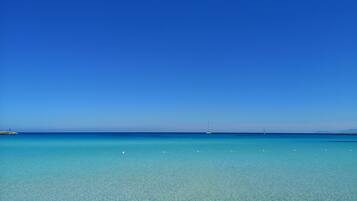 A clear blue sea with a distant sailboat and a rocky outcrop on the left.