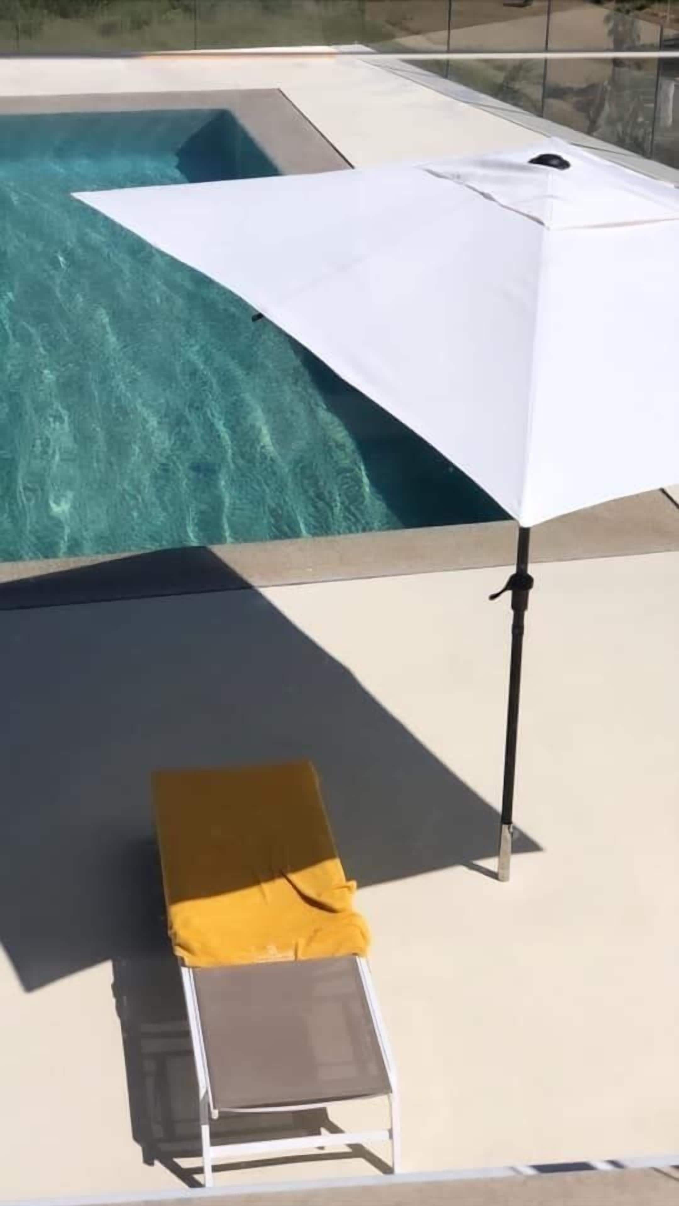 A poolside area with a white umbrella, a yellow cushion, and a lounge chair.