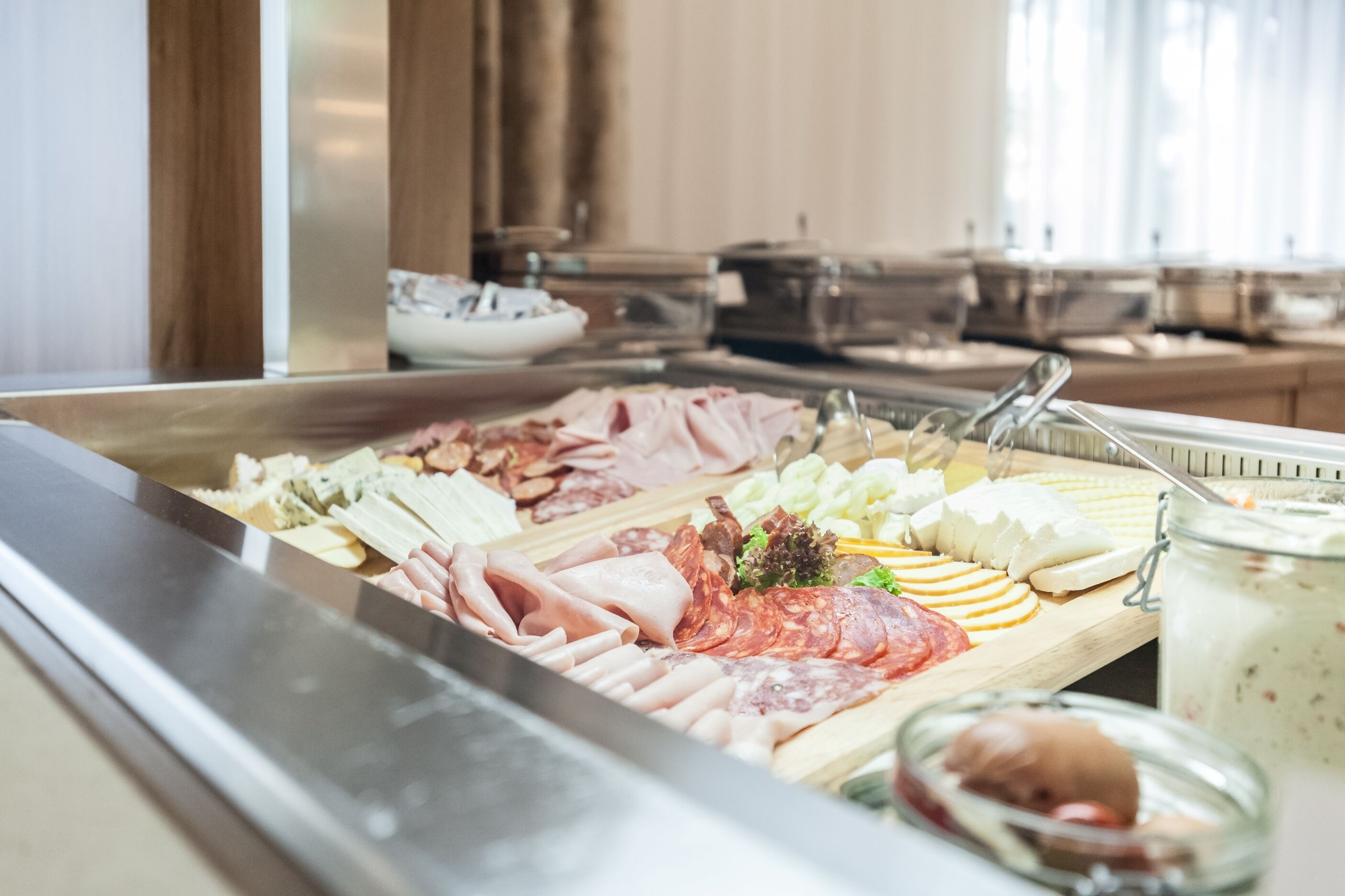 A buffet with various meats, cheeses, and a jar of yogurt parfait.