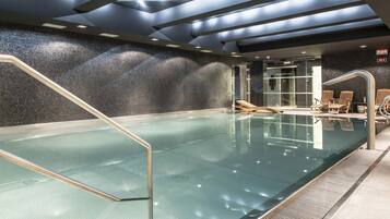 A modern indoor swimming pool with clear glass walls, a tiled wall, and a ceiling with recessed lighting.