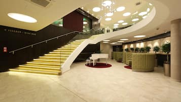 A modern lobby with a grand staircase, a piano, and a reception area with green seating.
