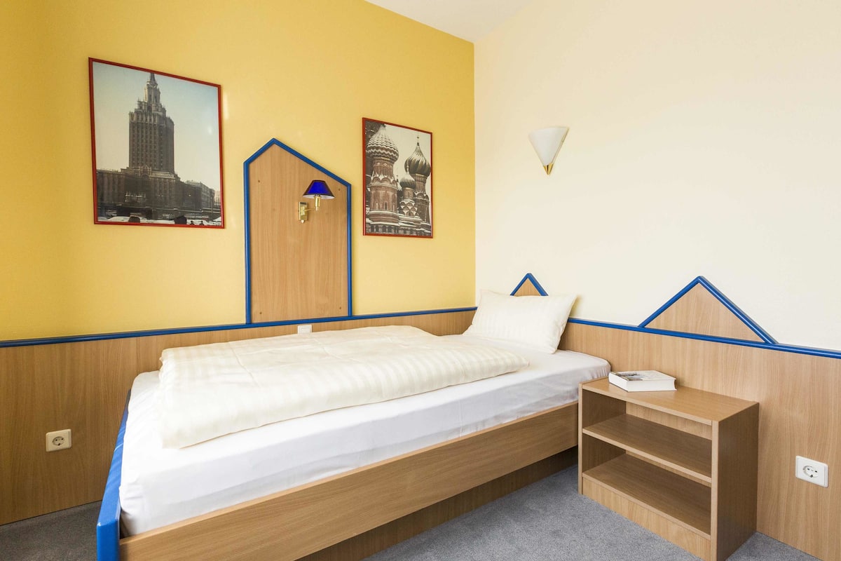 Habitación individual con una cama de madera, una mesita de noche pequeña y dos cuadros enmarcados en la pared.