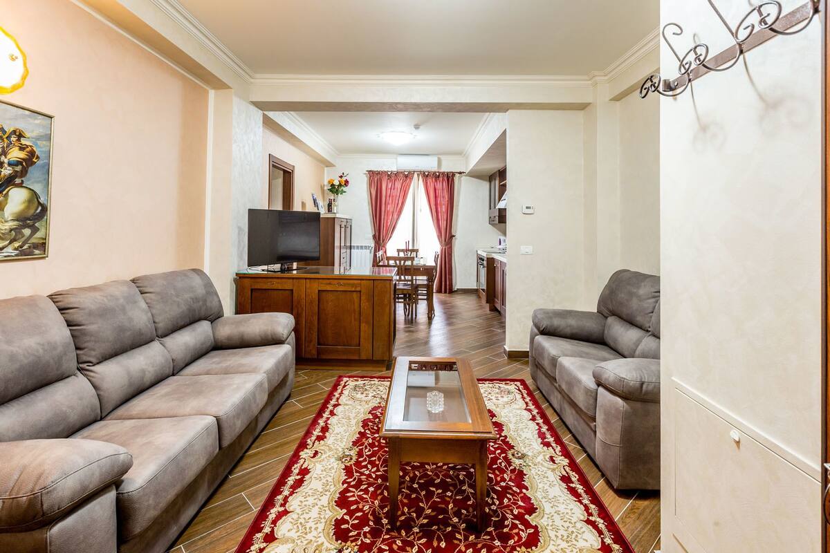 Ein Wohnzimmer mit zwei grauen Sofas, einem Couchtisch und einem Teppich in Rot und Weiß. Ein Fernseher steht auf einer Holzkommode, und im Hintergrund ist ein Essbereich mit Tisch und Stühlen zu sehen.