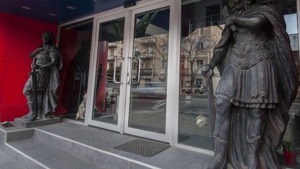 A building entrance with a glass door, a red wall, and two statues on the steps. - London Palace (Tbilisi)
