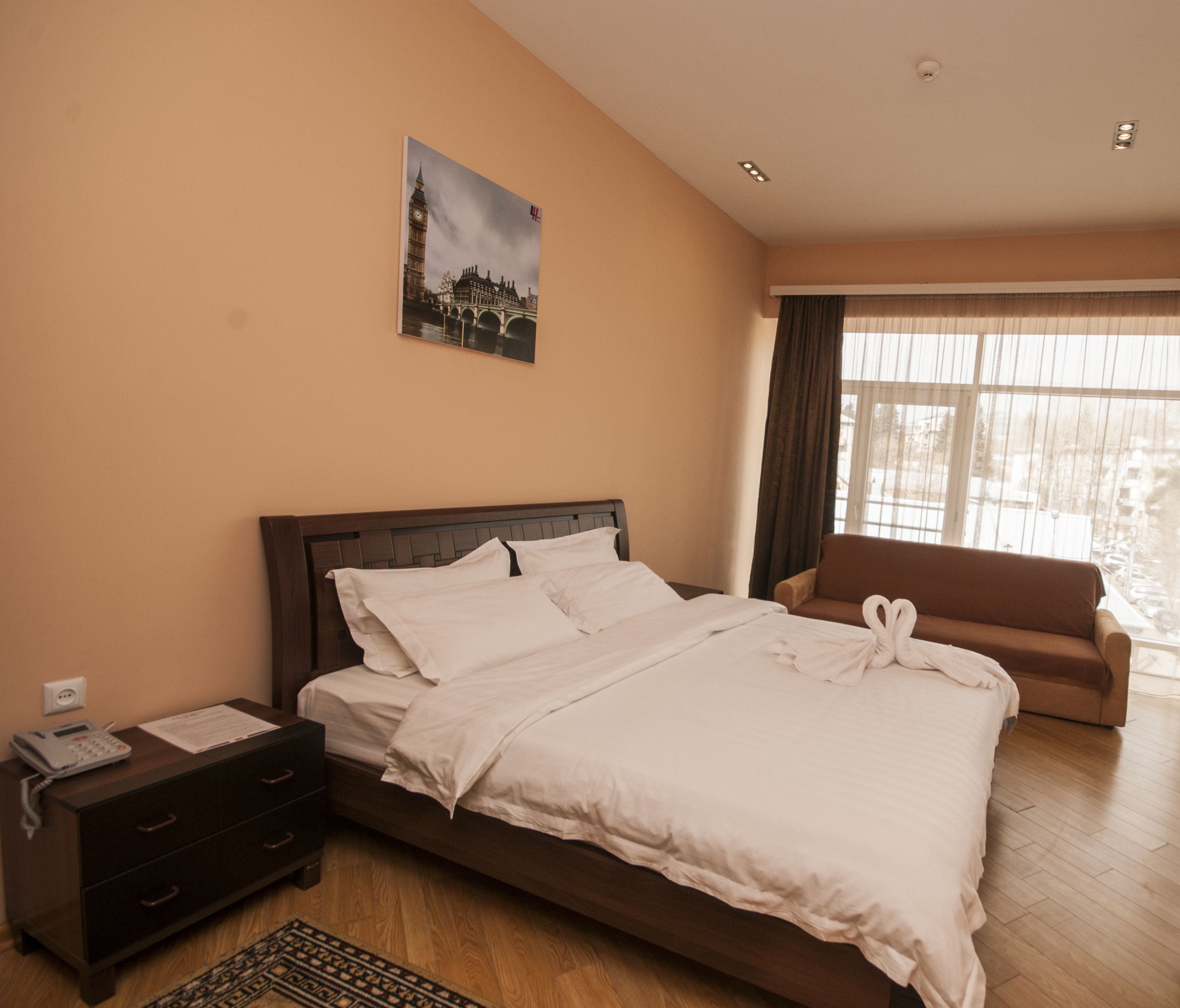 A neatly made bed with white linens, a wooden nightstand with a telephone, a brown sofa, and a large window with curtains.