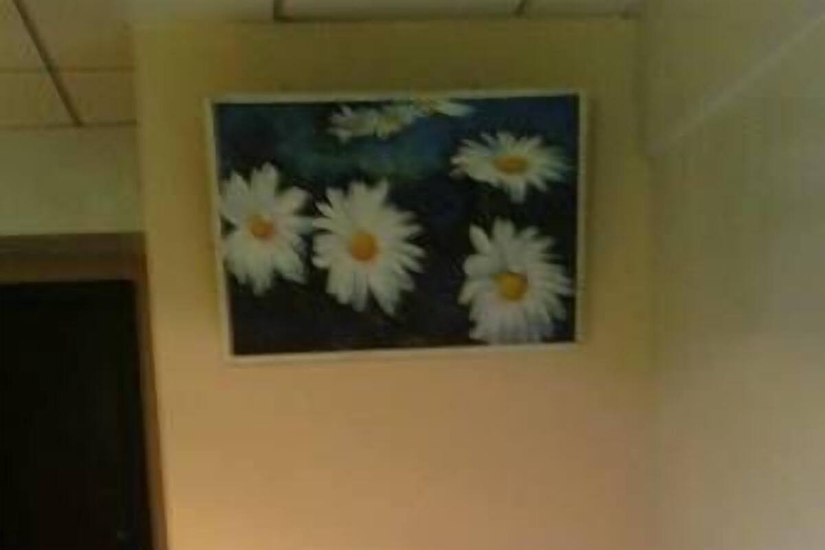 A neatly made bed with a white bedspread and pillows, a wooden headboard, and a painting of daisies on the wall above.