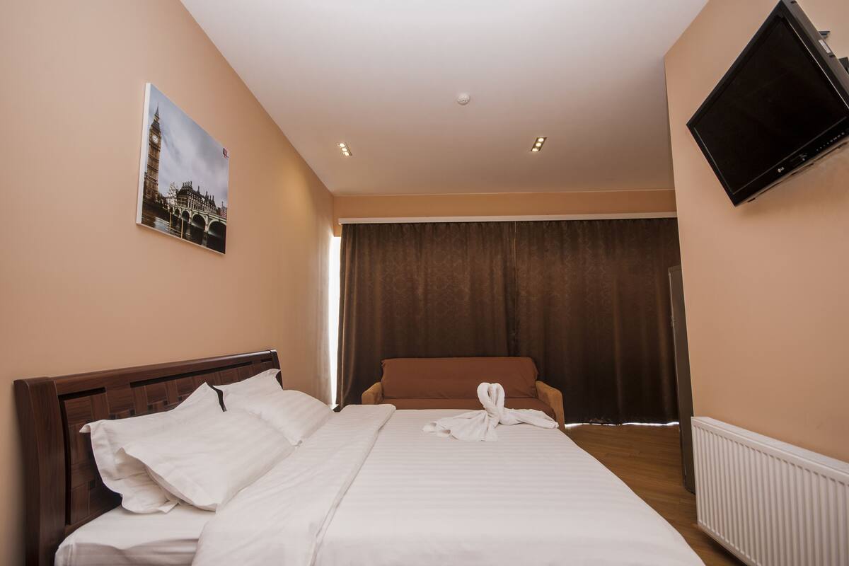 A hotel room with a bed, a flat-screen TV, a painting of a famous landmark, and a large window with curtains.