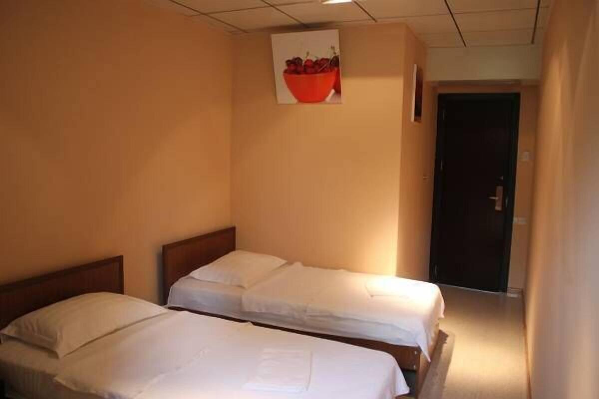A hotel room with two single beds, a wooden headboard, a wall-mounted picture of a red bowl, and a door leading to another room.