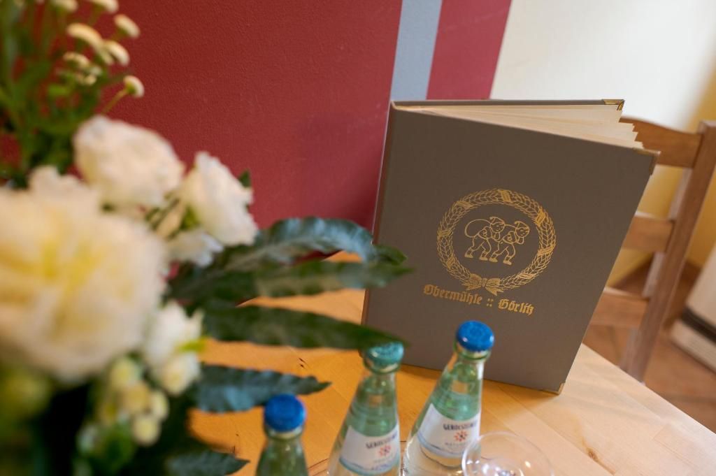 Un livre orné d’un emblème doré sur sa couverture, posé sur une table avec un vase de fleurs et trois bouteilles.