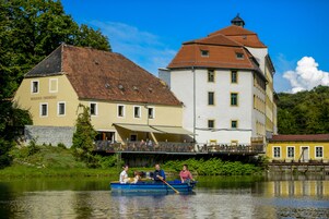 Ein Fluss mit einem Boot, das drei Personen und einen Hund befördert. Im Hintergrund sind zwei Gebäude in unterschiedlichem Baustil zu sehen.