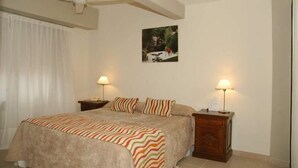 Habitación de hotel con una cama, dos mesitas de noche con lámparas, un ventilador de techo, un cuadro en la pared y una ventana con cortinas.
