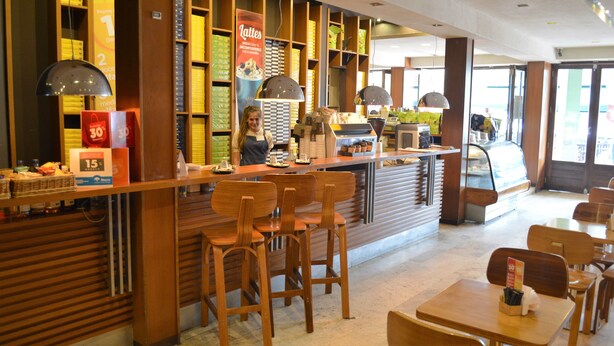 Un interior de café con muebles de madera, una barra con menú y un ventanal grande.