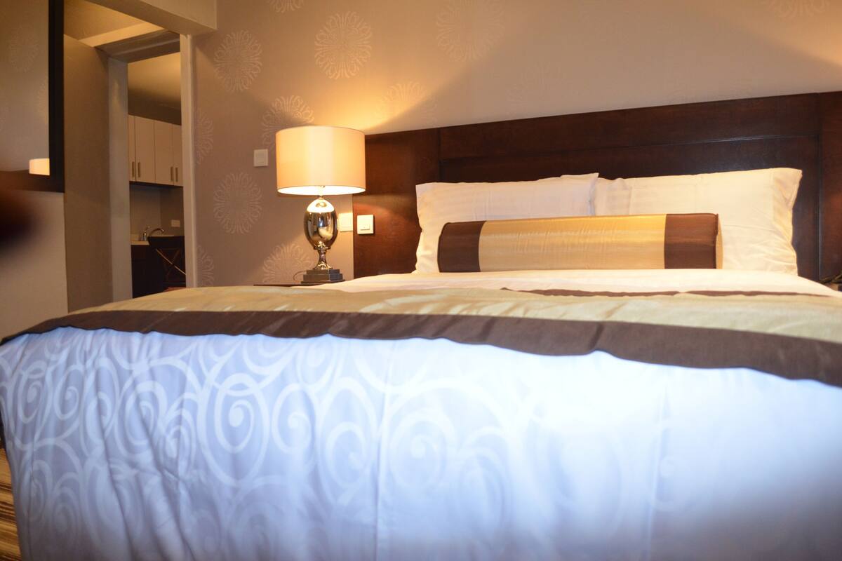 Une chambre d’hôtel avec un grand lit, une table de chevet avec une lampe et une vue sur un couloir menant à la salle de bain.
