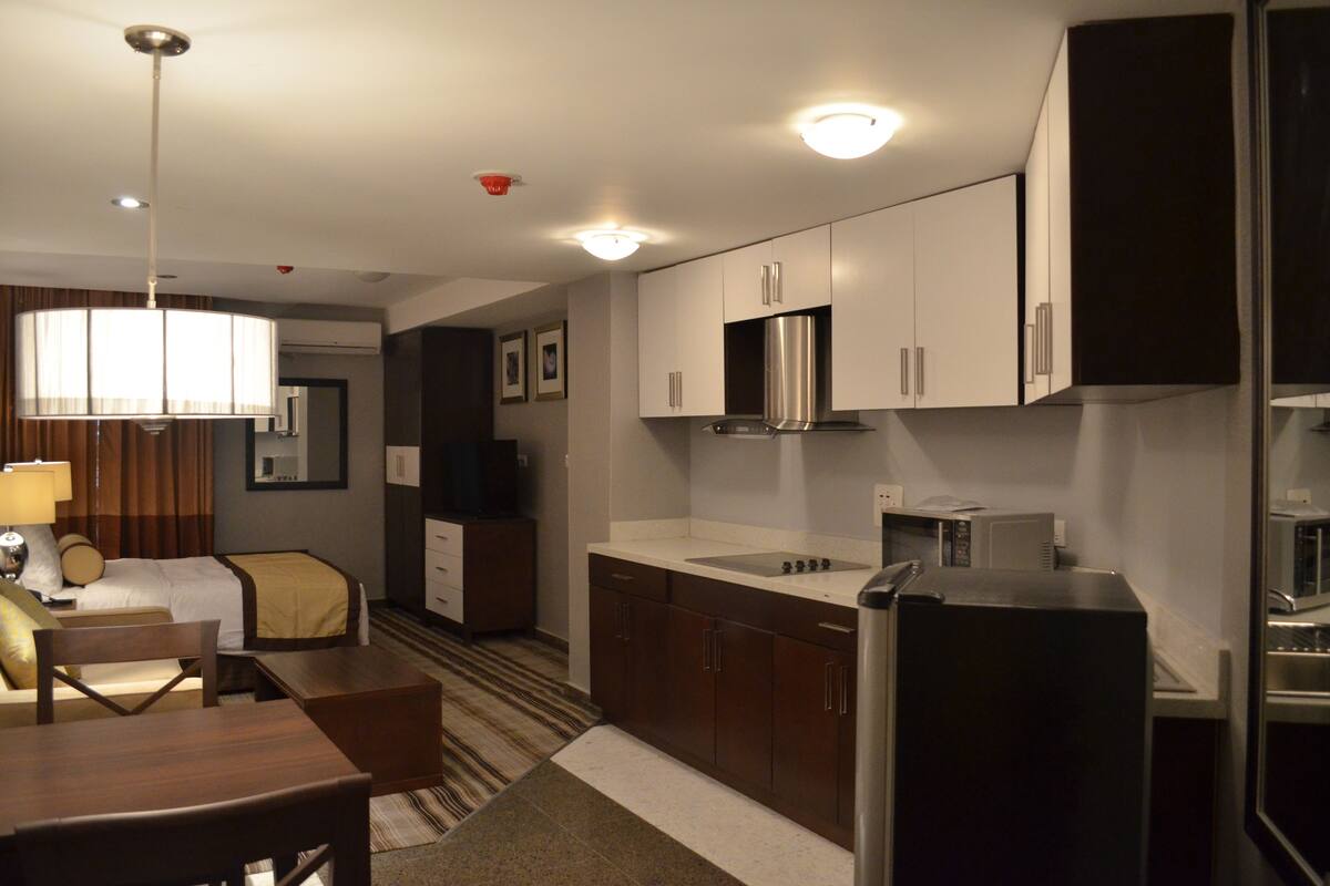 Une chambre d’hôtel comprenant un lit, un bureau, une chaise, une table à manger, un coin cuisine avec des placards et un réfrigérateur, ainsi qu’un luminaire suspendu.