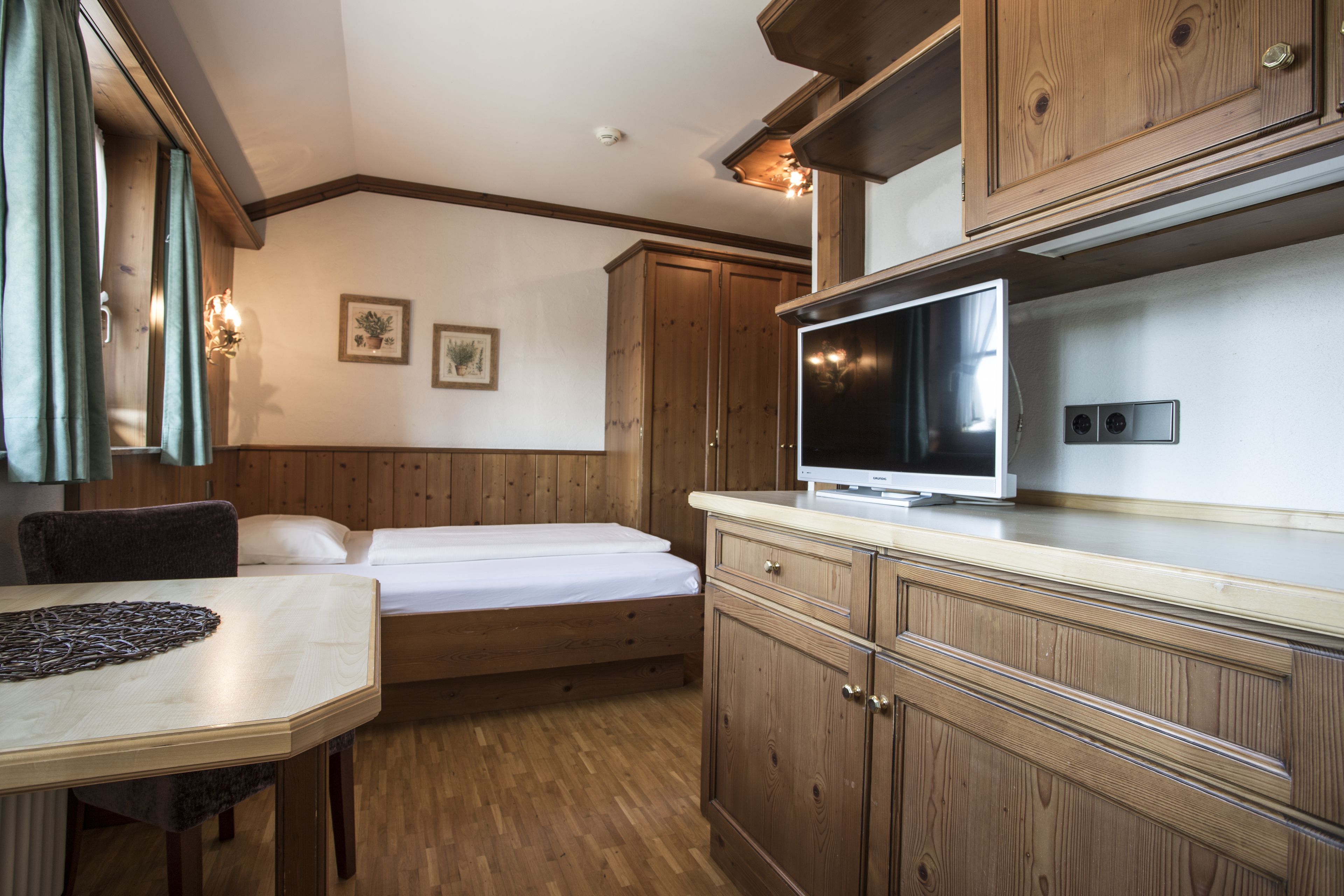 A compact room with a bed, a dining table, wooden cabinets, and a television.