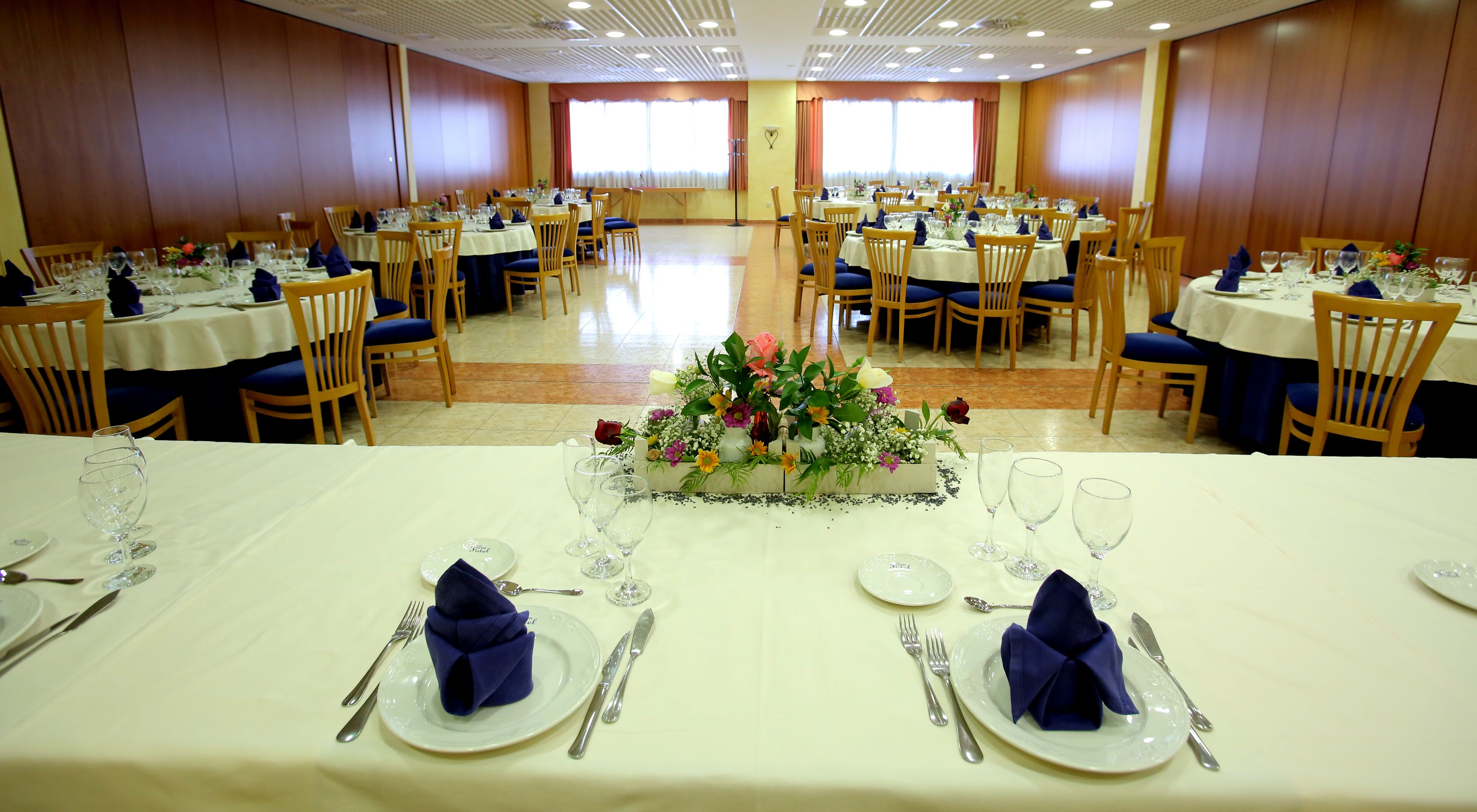 Un salón de banquetes con varias mesas redondas, cada una con mantel blanco, servilletas azules y centros de mesa florales.