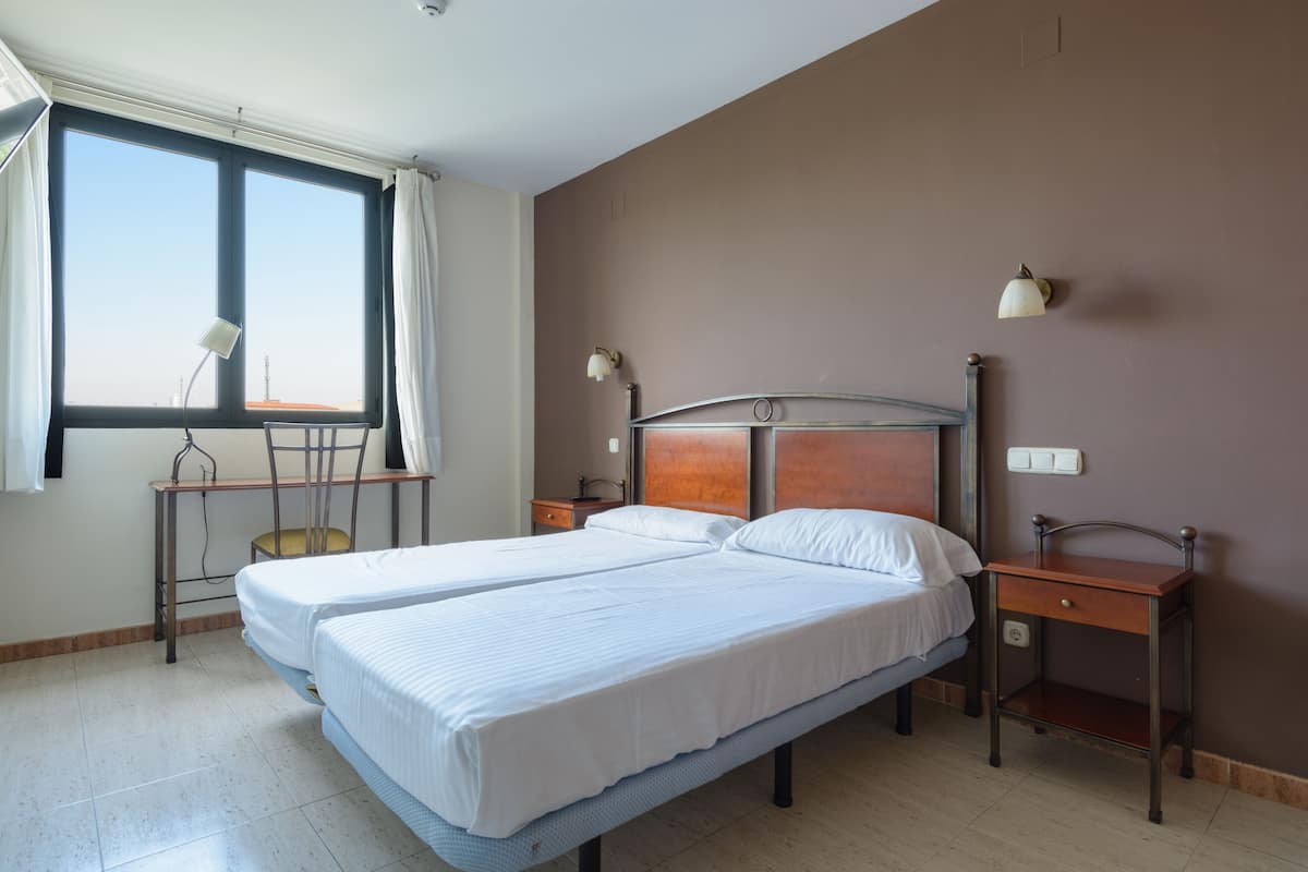 Una cama doble con cabecera de madera, dos mesitas de noche, un escritorio con silla y una ventana con cortinas.