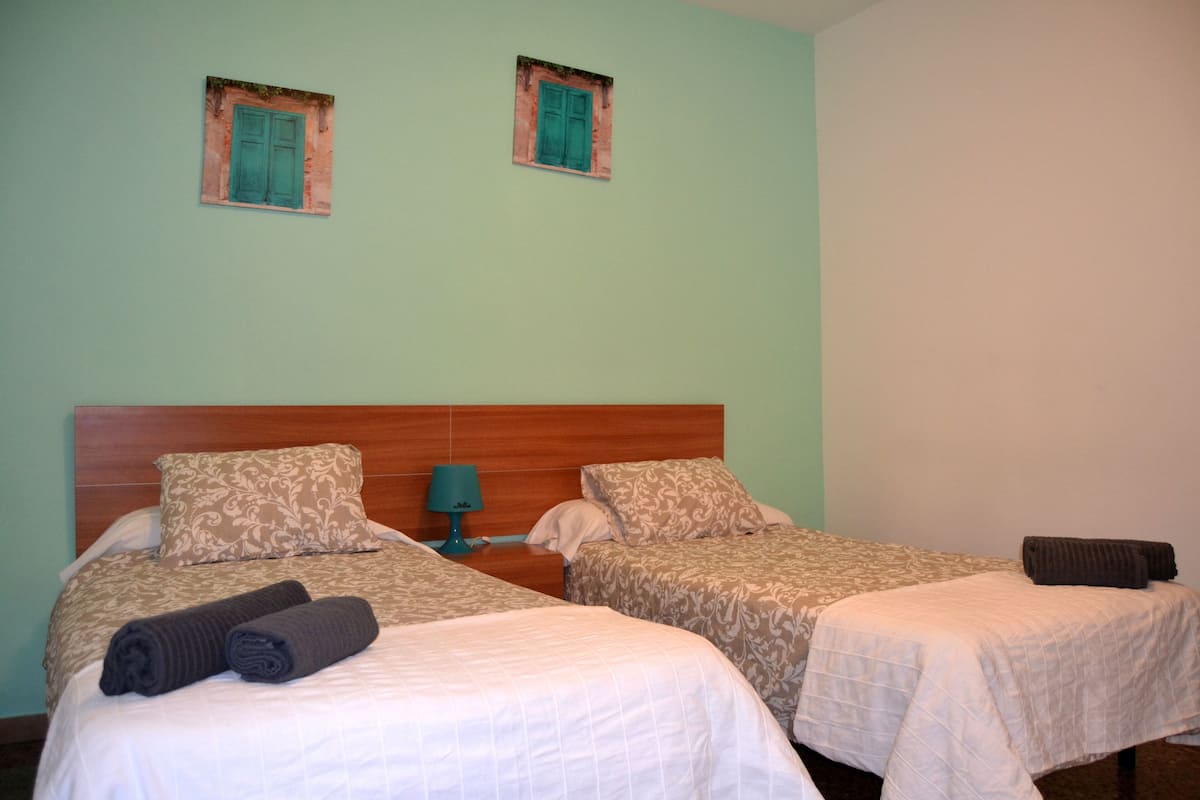 A hotel room with two single beds, a wooden headboard, a nightstand with a lamp, and two small windows with green shutters.