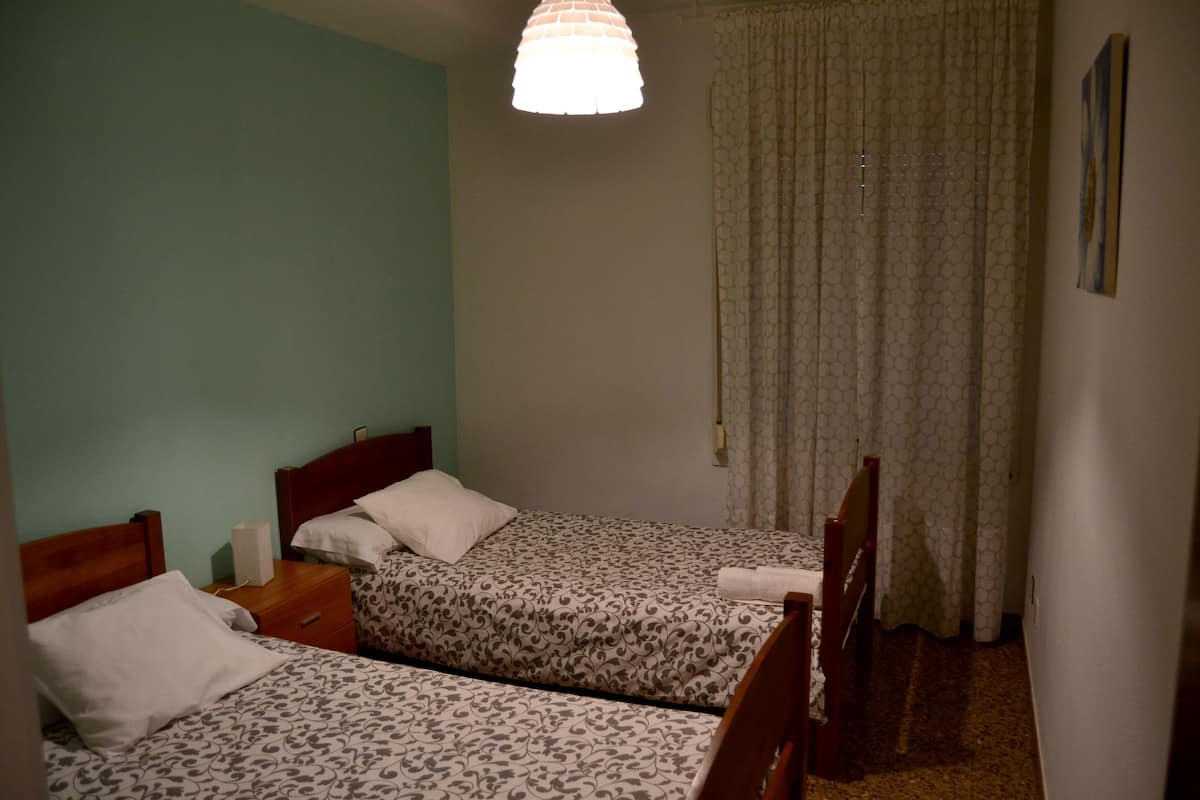 A room with two beds, a wooden headboard, a bedside table, a lamp, and a curtain.