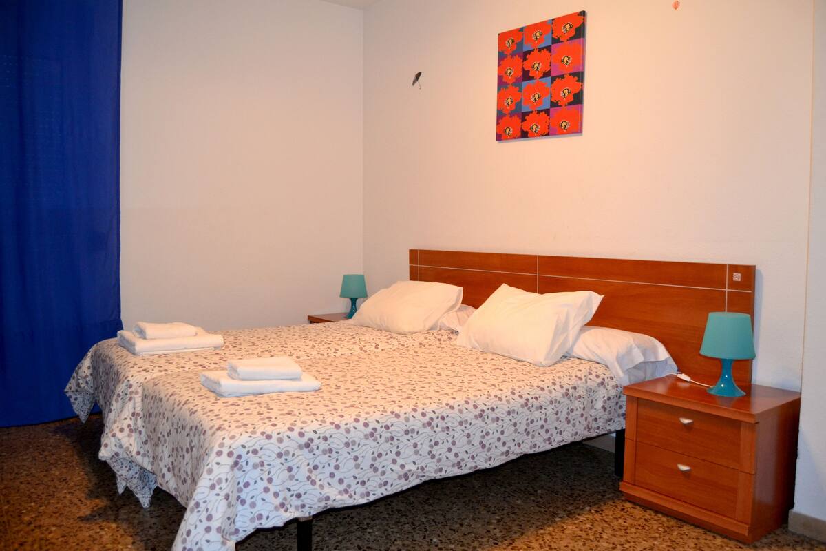 A neatly made bed with a floral bedspread, two pillows, and folded towels. There is a wooden headboard, a nightstand with a lamp, and a wall art piece above the bed.