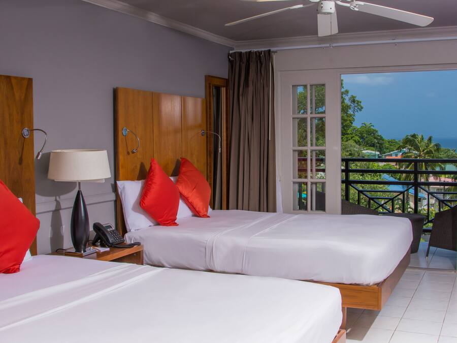A hotel room with two beds, a ceiling fan, a window with a view of the ocean, and a balcony with a chair.