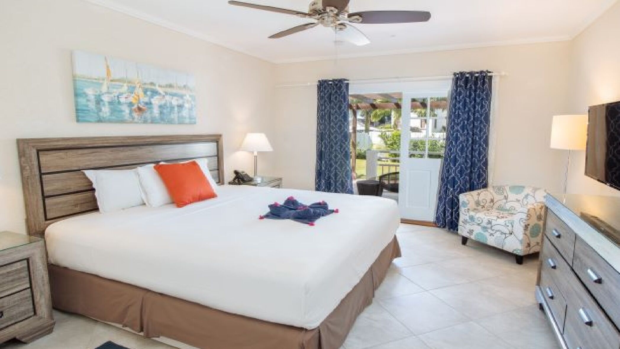 A hotel room with a bed, a ceiling fan, a painting, a TV, and a view of the outdoors.