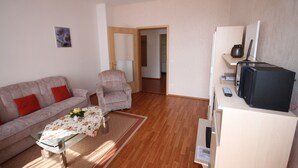 Een woonkamer met een bank, fauteuil en een tafel met een vaas. Er staat een televisie op een dressoir en een deur die naar een andere kamer leidt.