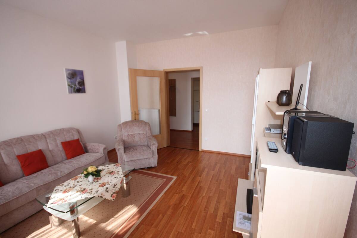 Een woonkamer met een bank, fauteuil en een tafel met een vaas. Er staat een televisie op een dressoir en een deur die naar een andere kamer leidt.