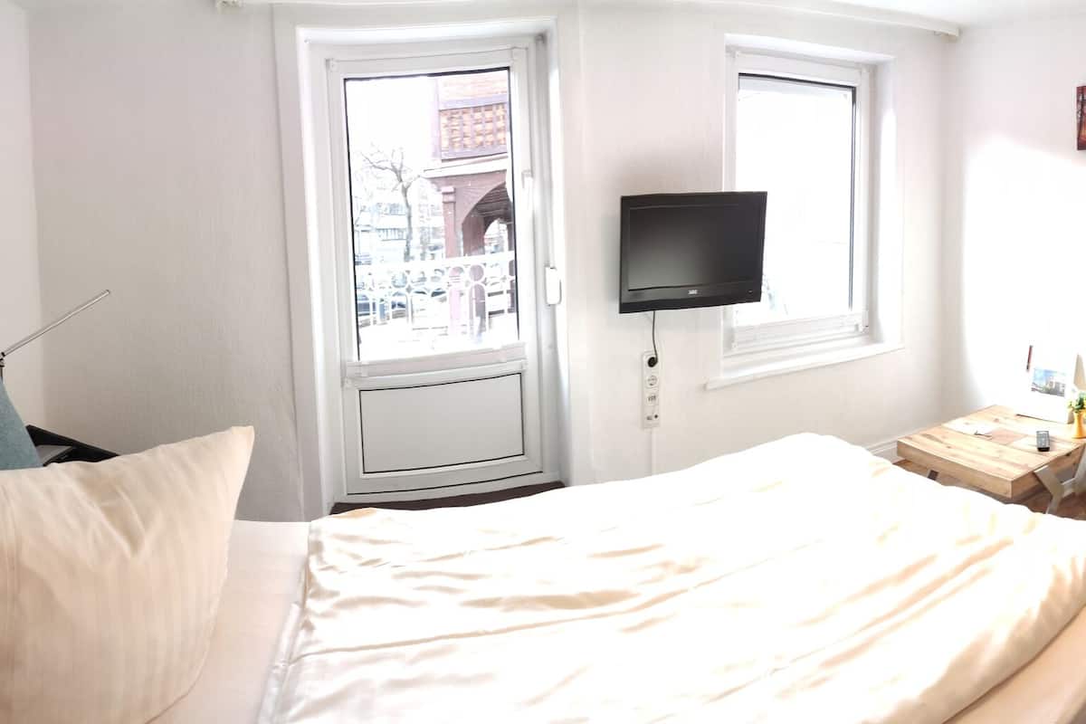 Een slaapkamer met een bed, een grijze fauteuil, een televisie, een raam en een deur.