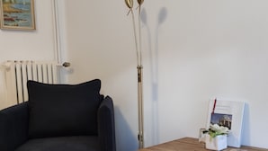Een kamer met een zwarte bank, een houten tafel, een vloerlamp, een radiator en een lijst met een afbeelding aan de muur.