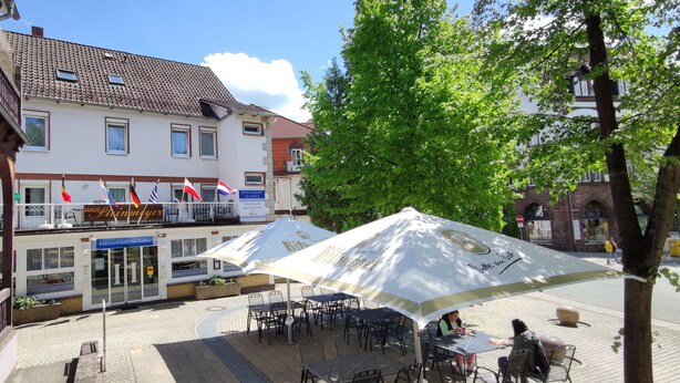 Een restaurant met terras, een gebouw met meerdere vlaggen, en een boom op de voorgrond.