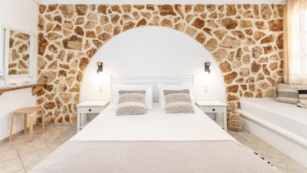 A bedroom with a stone wall, a bed with patterned pillows, a bedside table, a small stool, and a woven basket. - Studios Tasia (Naxos)