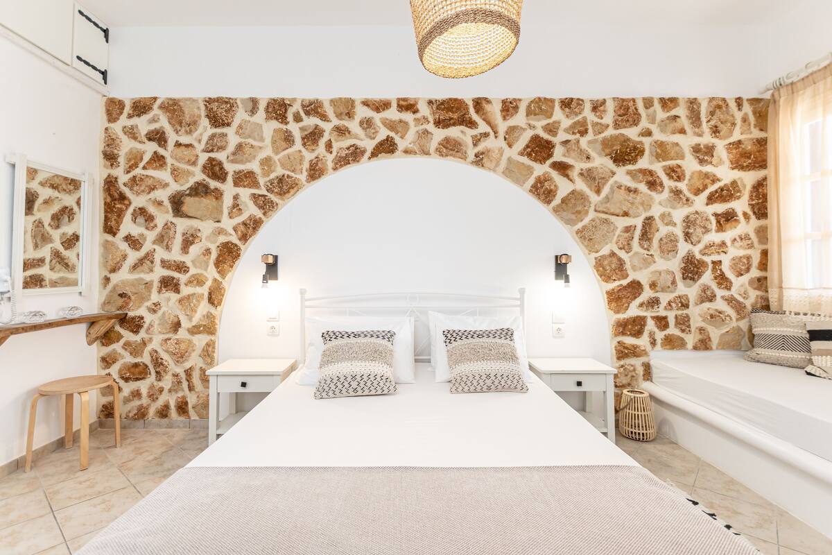 A bedroom with a stone wall, a bed with patterned pillows, a bedside table, a small stool, and a woven basket.