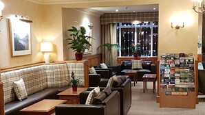 A hotel lobby with a sofa, armchairs, a coffee table, a plant, a window with curtains, and a display of magazines. - Loch Long Hotel (Arrochar)