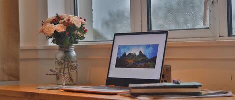 A wooden desk with a laptop, a framed picture, a vase with flowers, and a notebook.