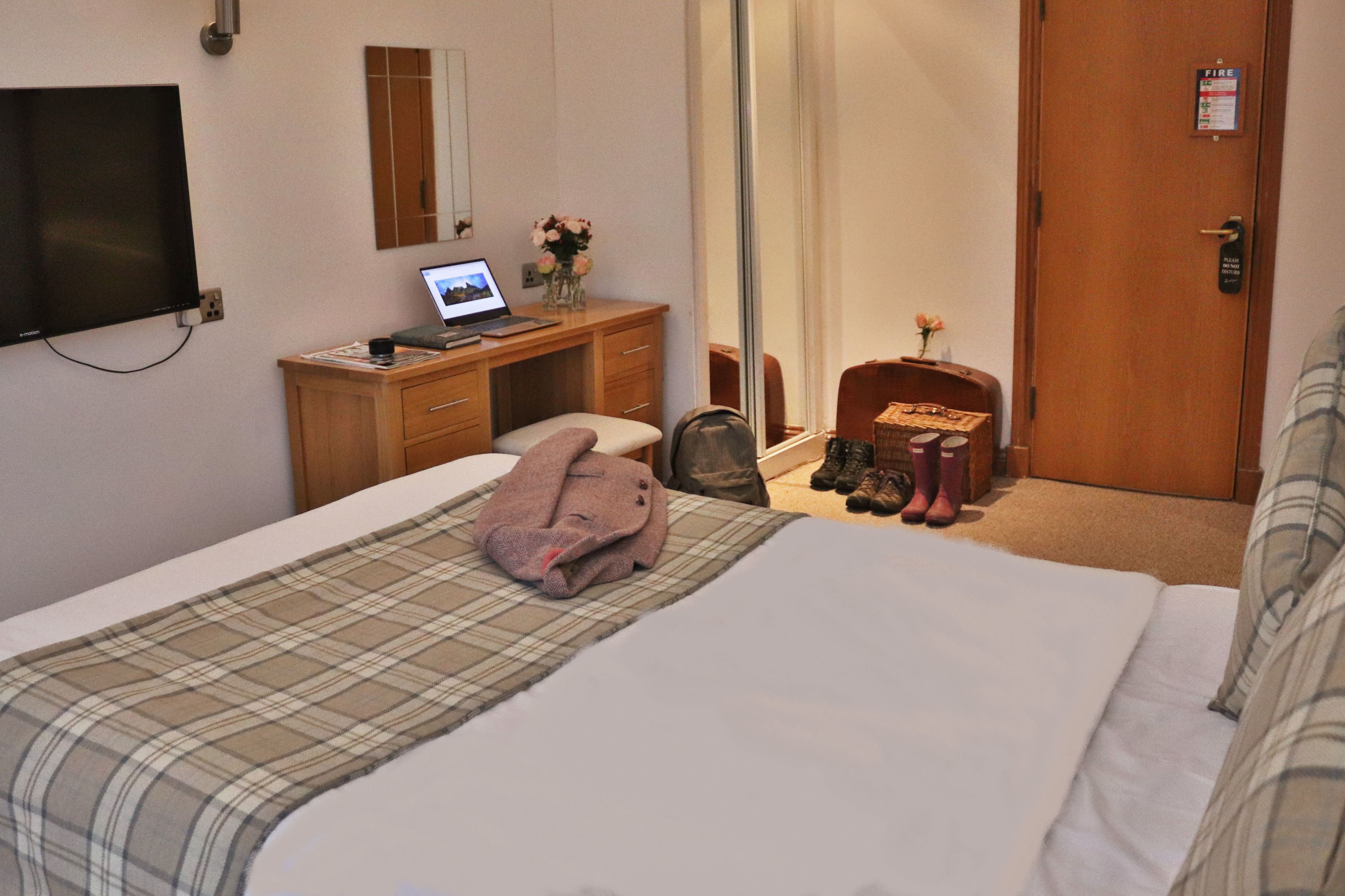 A hotel room with a bed, a desk with a laptop, a mirror, a wardrobe, and a door with a fire alarm.