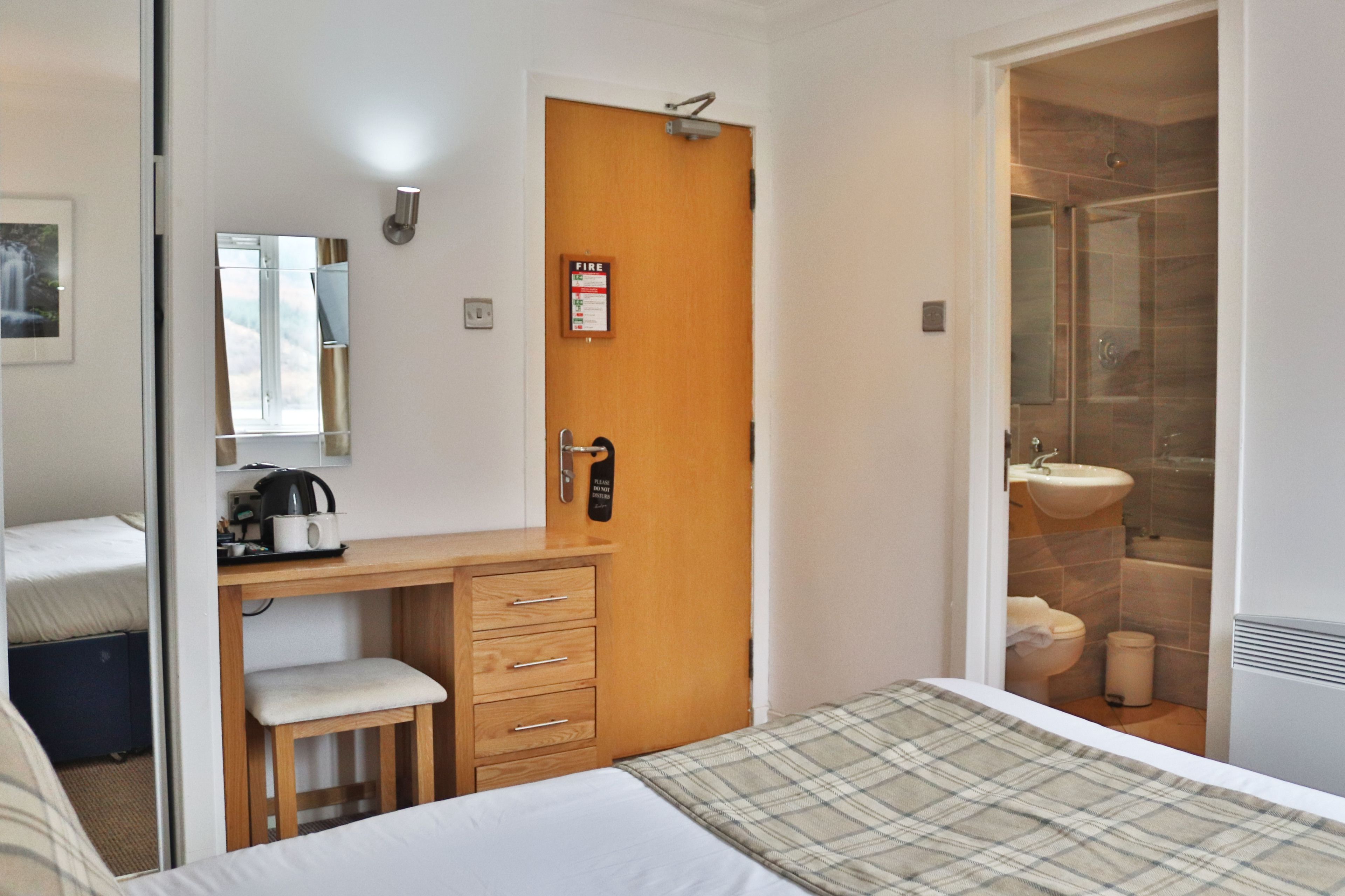 Habitación de hotel con cama, escritorio de madera con cajones, silla, espejo, puerta con señal de seguridad contra incendios y baño con lavamanos y retrete.