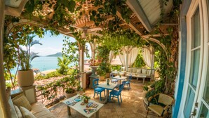 Un patio cubierto con vista al mar, que incluye un sofá, sillas, una mesa con revistas y plantas en macetas.