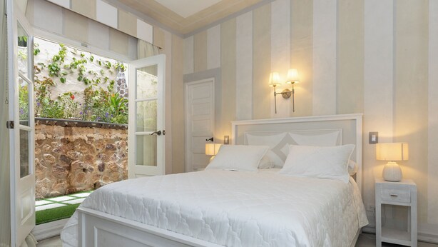 A bedroom with a bed, a nightstand, a wall-mounted lamp, and a door leading to a garden with plants. - Villa Caprichosa   (Taboga)