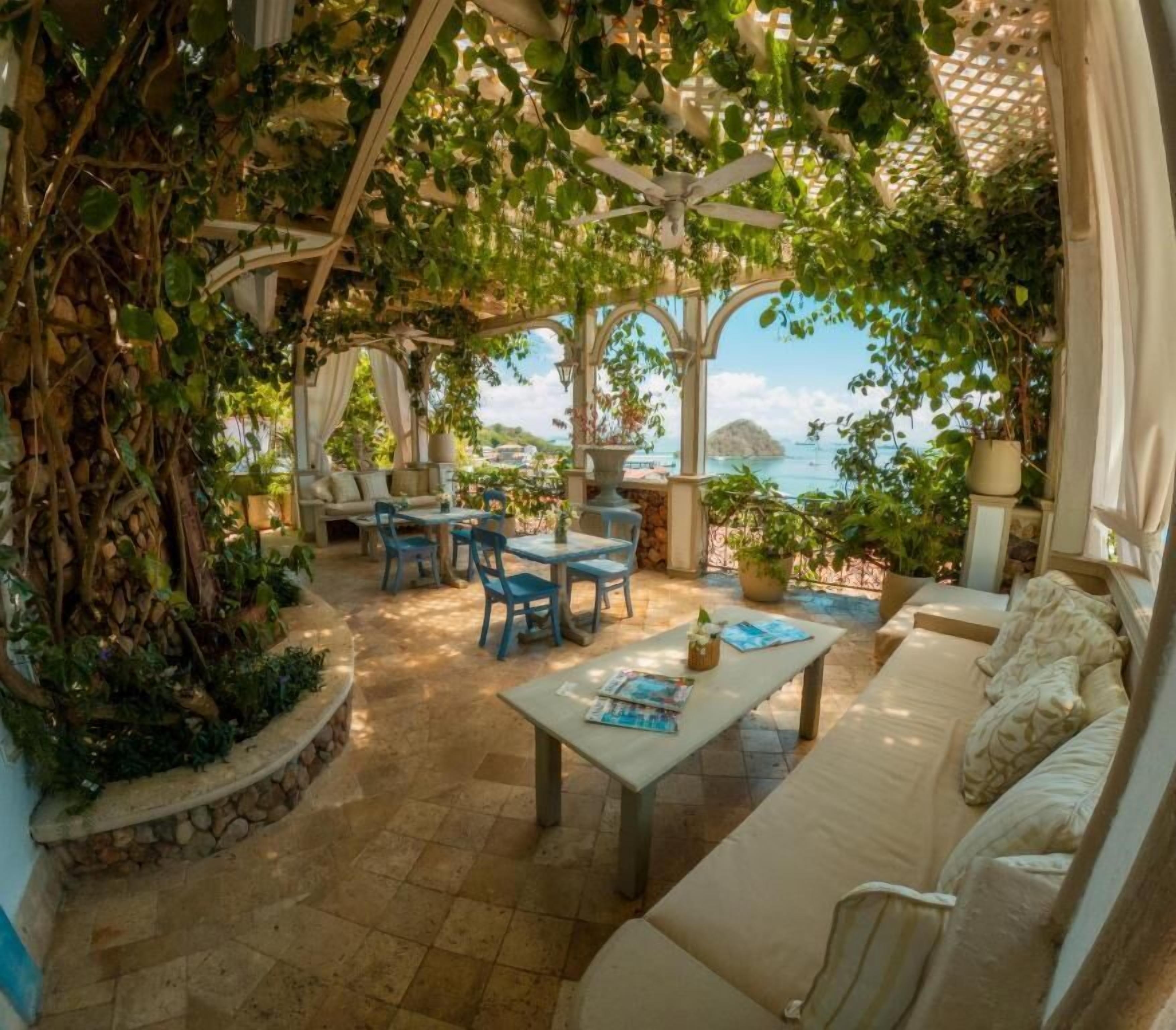 A covered outdoor seating area with a view of the sea, stone flooring, and potted plants.