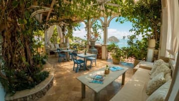 A covered outdoor seating area with a view of the sea, stone flooring, and potted plants.