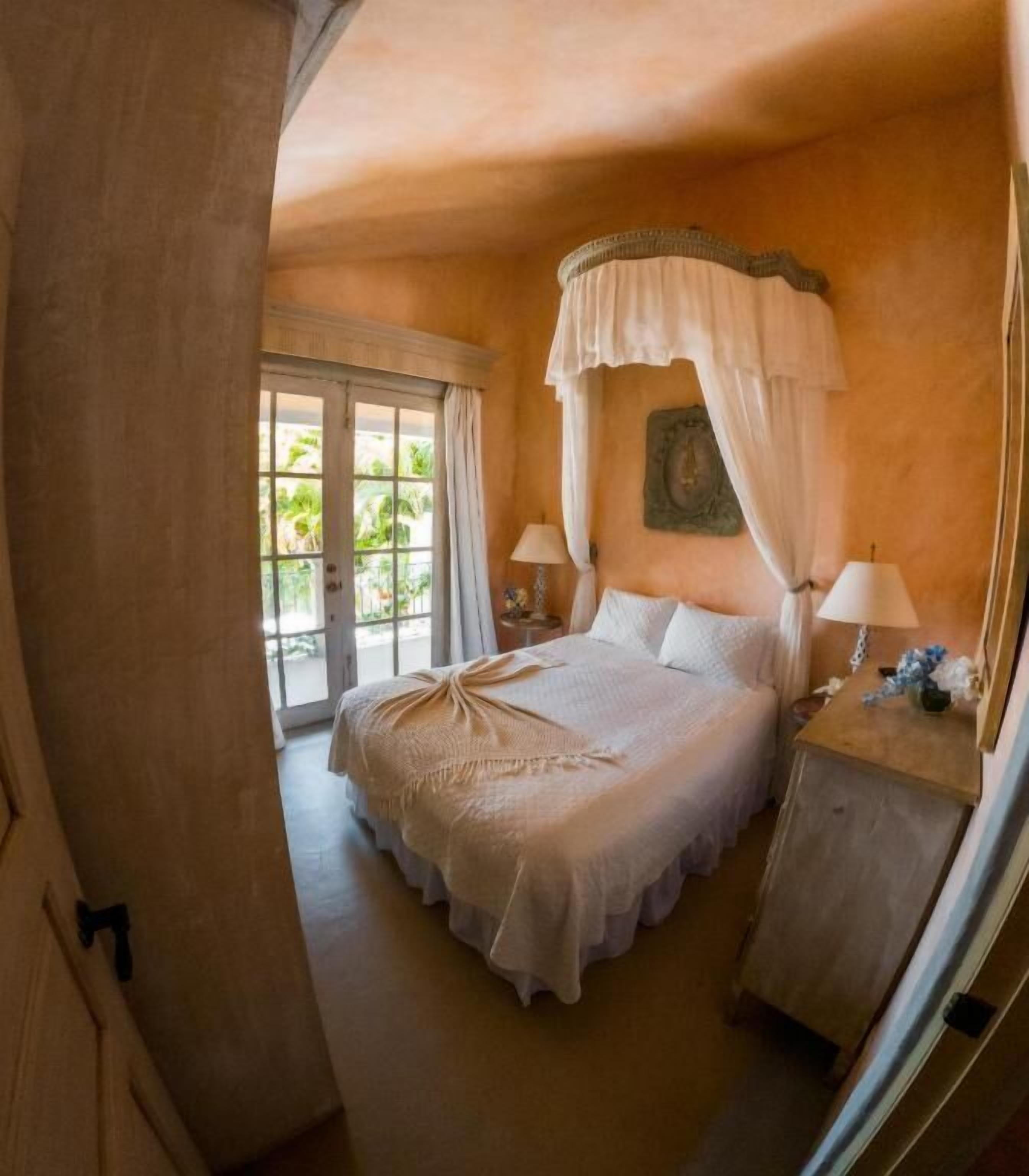 A bedroom with a canopy bed, two bedside tables with lamps, a large window with curtains, and a view of greenery outside.