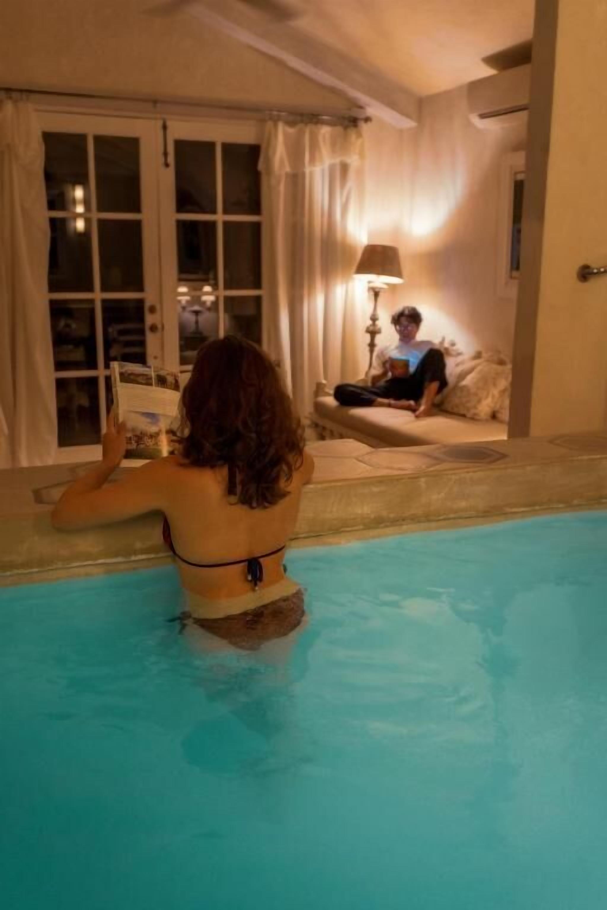 Una persona sentada en una piscina leyendo un libro, otra persona sentada en un sofá al fondo, y una lámpara sobre la mesa.
