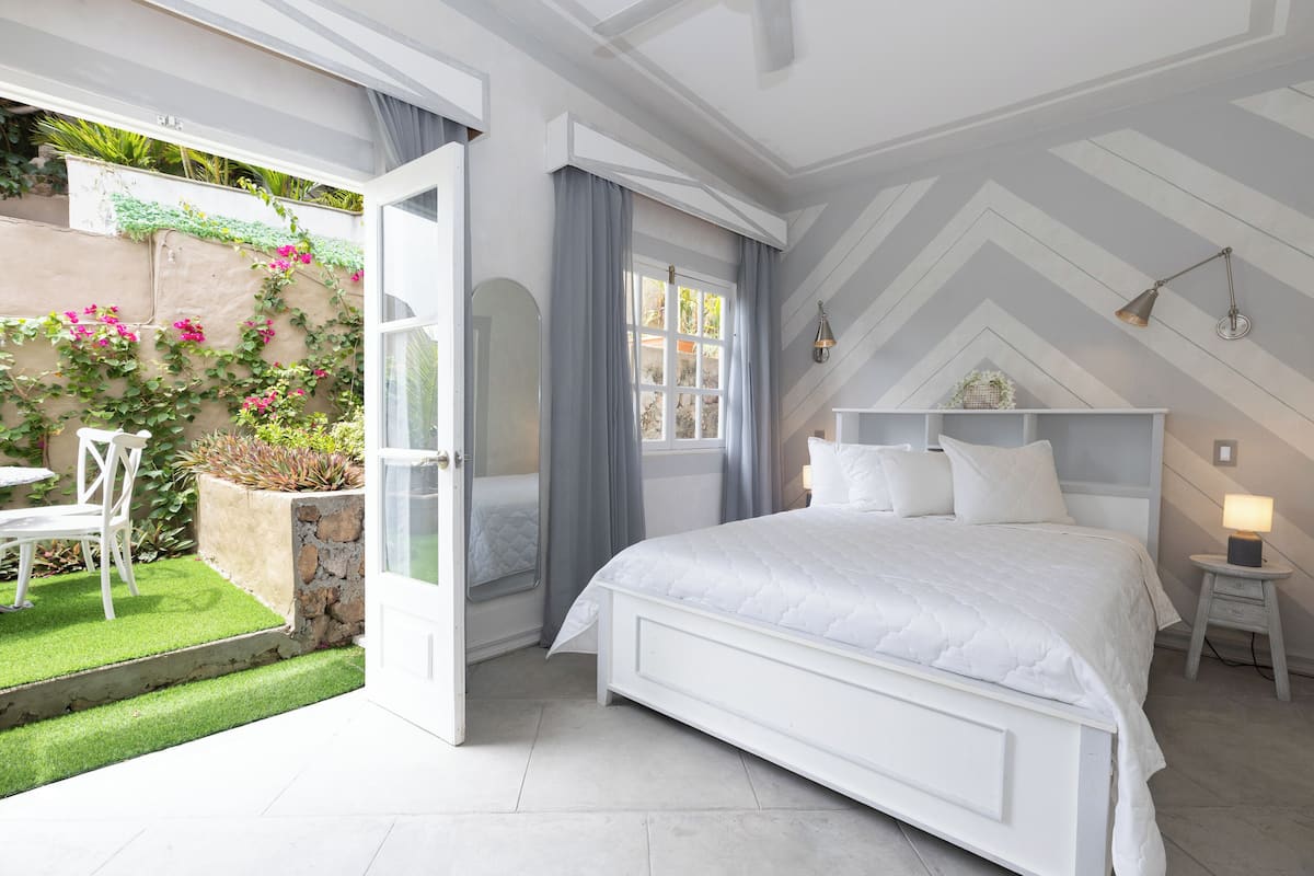 Ein Schlafzimmer mit einem großen Bett, einem Nachttisch mit Lampe und Blick auf einen Garten durch eine geöffnete Tür.