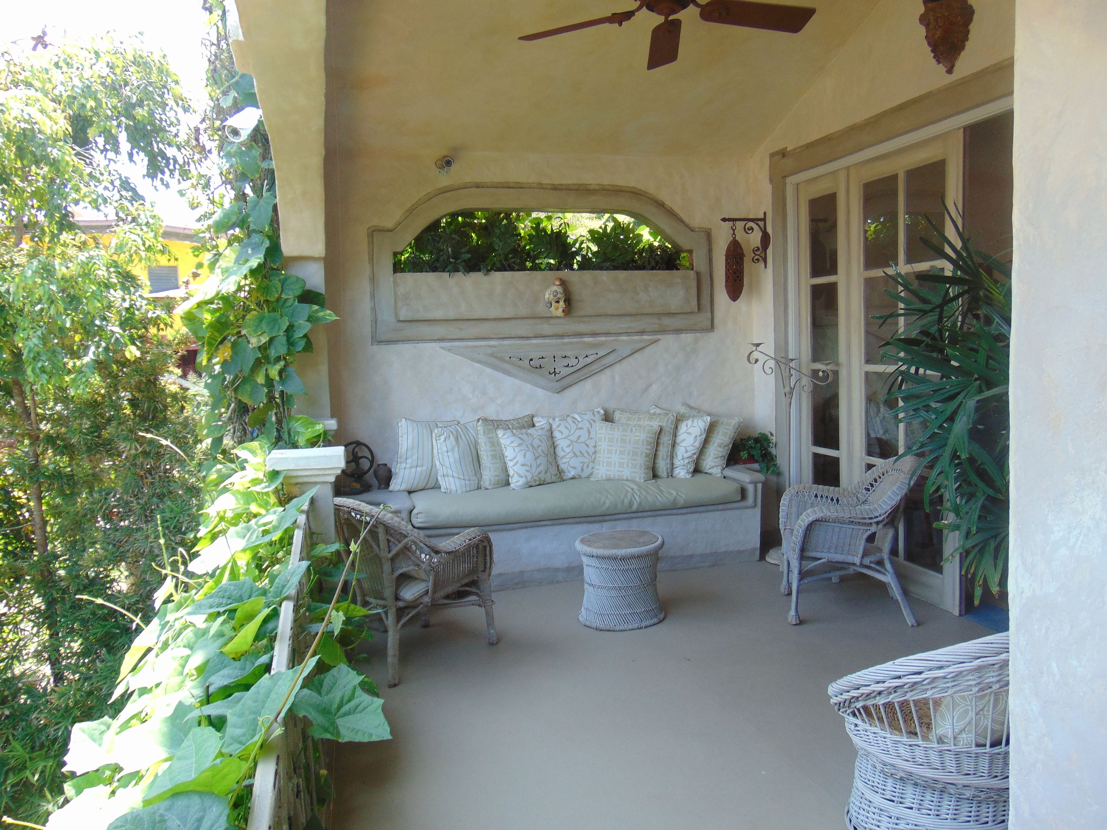 A sunlit patio with a cushioned seating area, wicker chairs, and a ceiling fan.