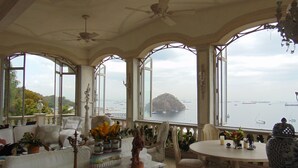 Amplia habitación con techo alto, ventanas grandes que ofrecen vista al mar y una isla distante, una mesa redonda con sillas y un sofá con adornos.