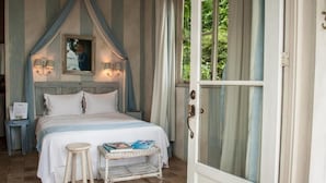 Un dormitorio con cama, mesita de noche y un pequeño taburete. Se observa una puerta con panel de vidrio y cortinas.