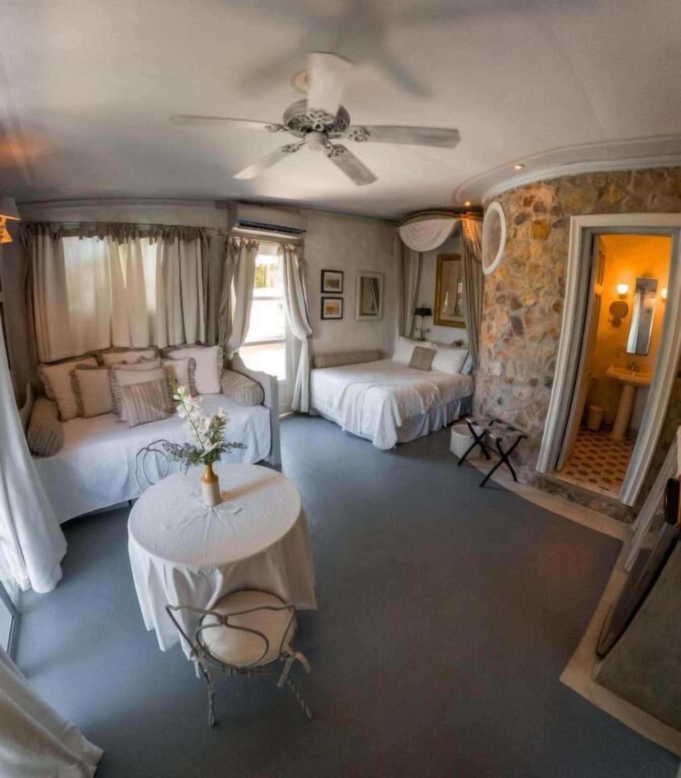 A hotel room with a bed, a smaller bed, a round table with a chair, and a ceiling fan.