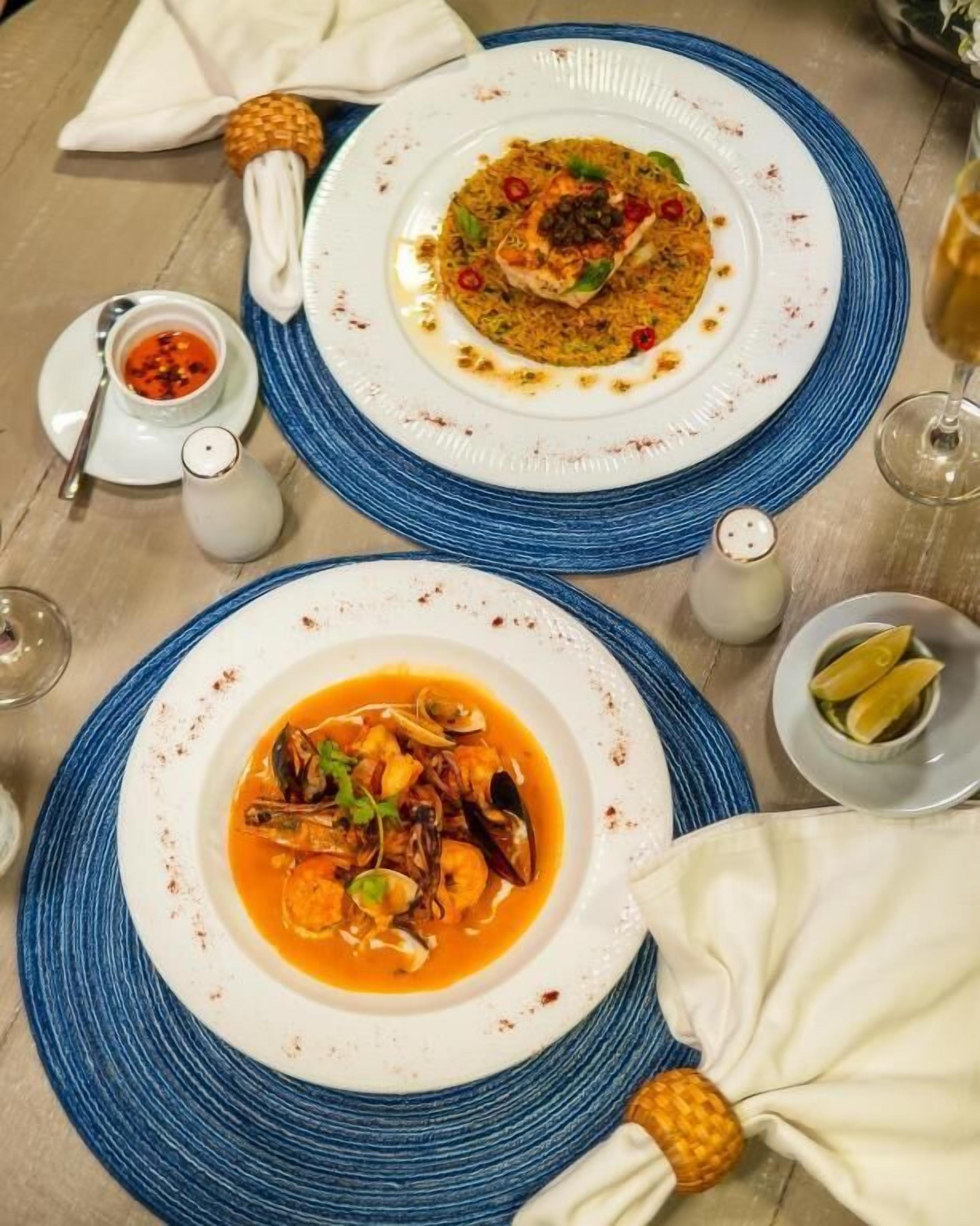 Duas travessas de pratos de frutos do mar sobre uma mesa com jogos americanos azuis, um saleiro e uma pequena tigela com gomos de limão.